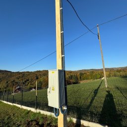 Instalatorstwo Elektryczne Kacper Szymala - Nowo zainstalowana skrzynka elektryczna na drewnianym słupie, z niebieskimi rurami karbowanymi, na tle krajobrazu z lasem i niebieskim niebem.