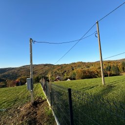 Instalatorstwo Elektryczne Kacper Szymala - Słup energetyczny z przyłączem kablowym i skrzynką licznikową na tle krajobrazu z ogrodzeniem panelowym.