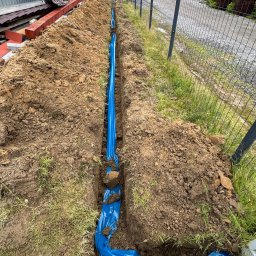 Instalatorstwo Elektryczne Kacper Szymala - Wykop z niebieską folią ochronną w ziemi, prawdopodobnie pod instalację. Widoczny fragment ogrodzenia i trawa. Wykop świeżo zrobiony, ziemia wokół.