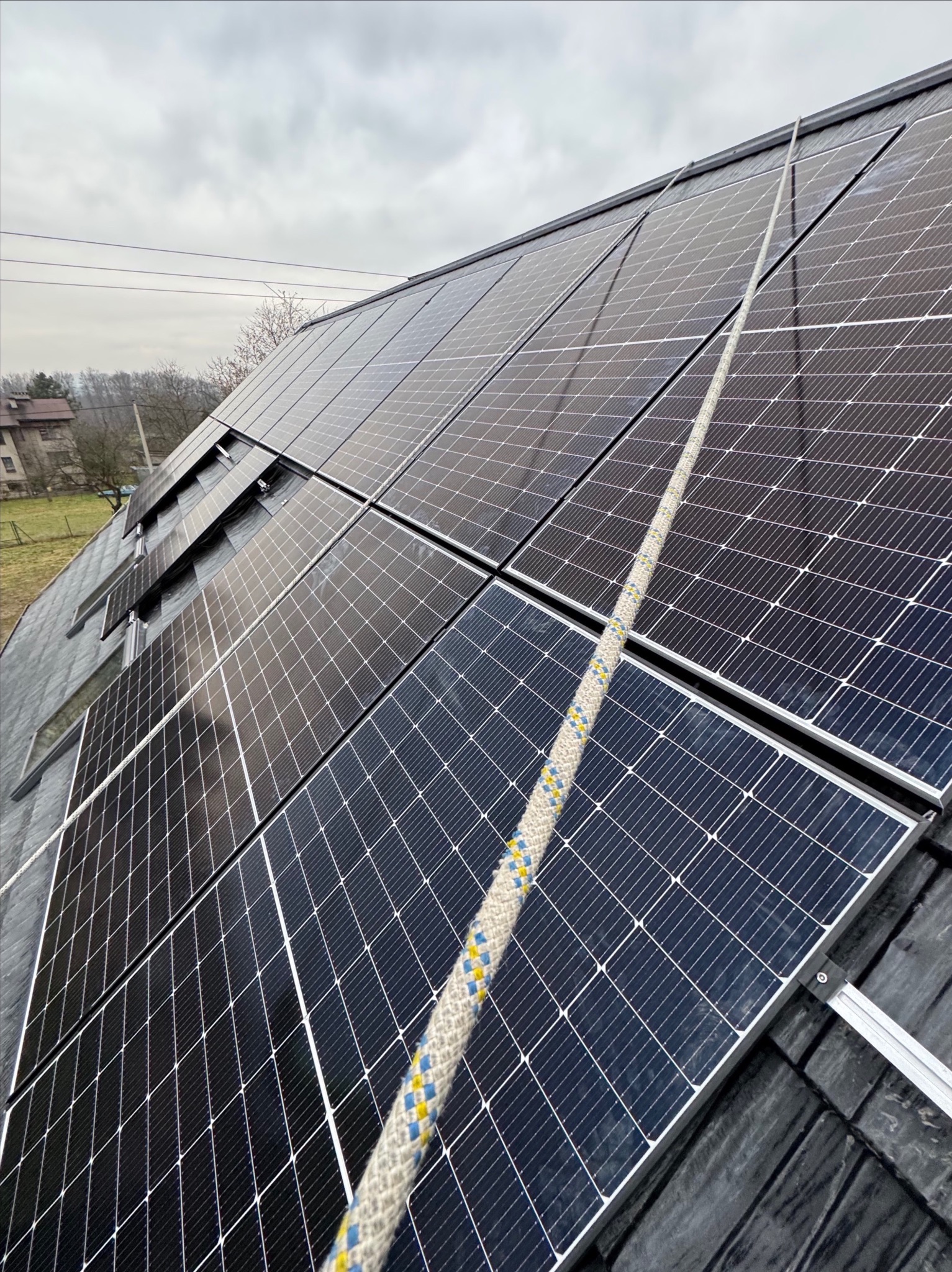 Panele słoneczne na dachu z widoczną liną asekuracyjną, montaż na czarnej papie, pochmurne niebo w tle.