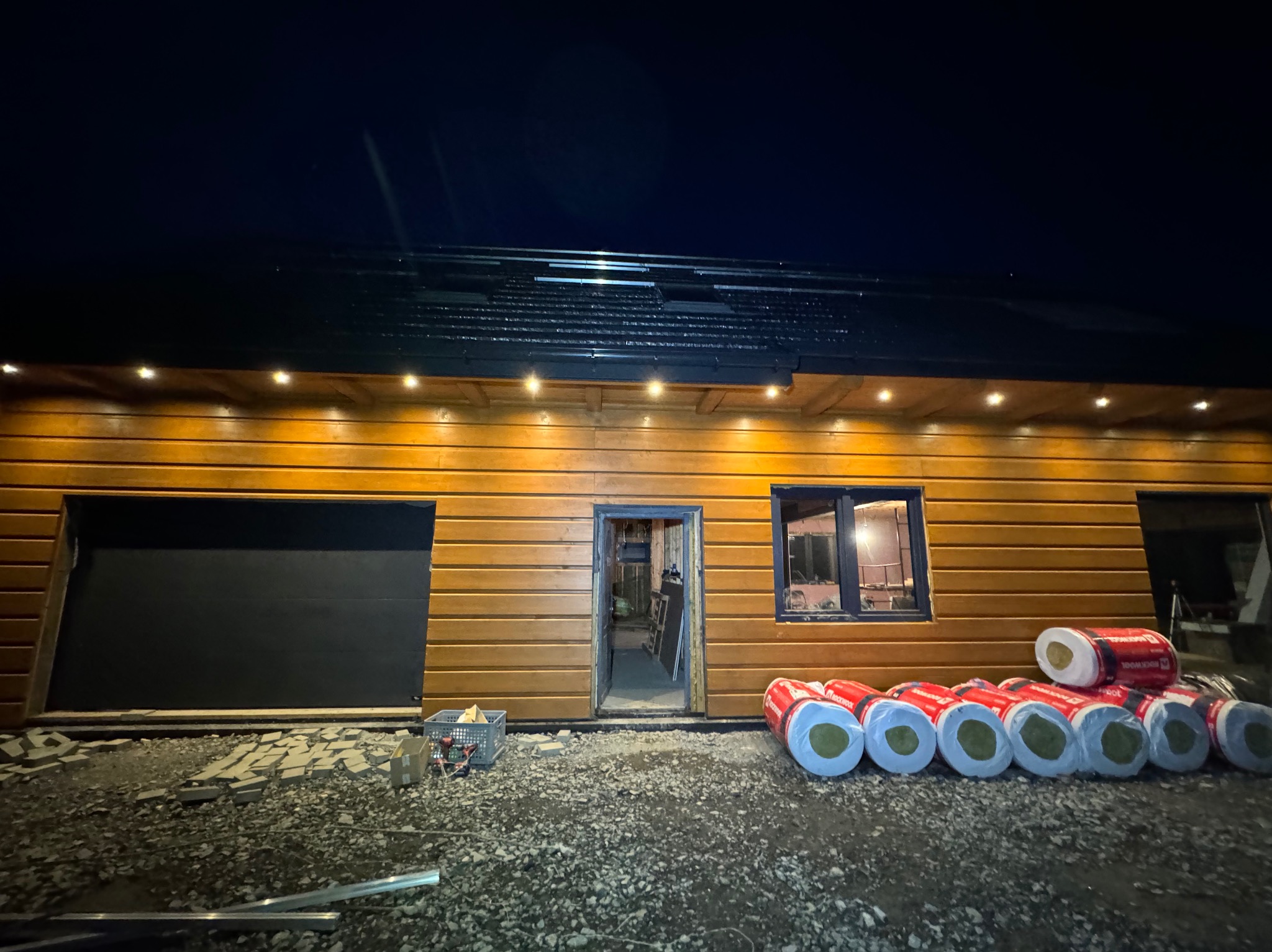 Drewniany budynek z oknem i otwartymi drzwiami, oświetlony punktowymi lampkami na zewnątrz, z materiałami izolacyjnymi Rockwool ułożonymi przed budynkiem, widoczne narzędzia w koszyku i elementy...