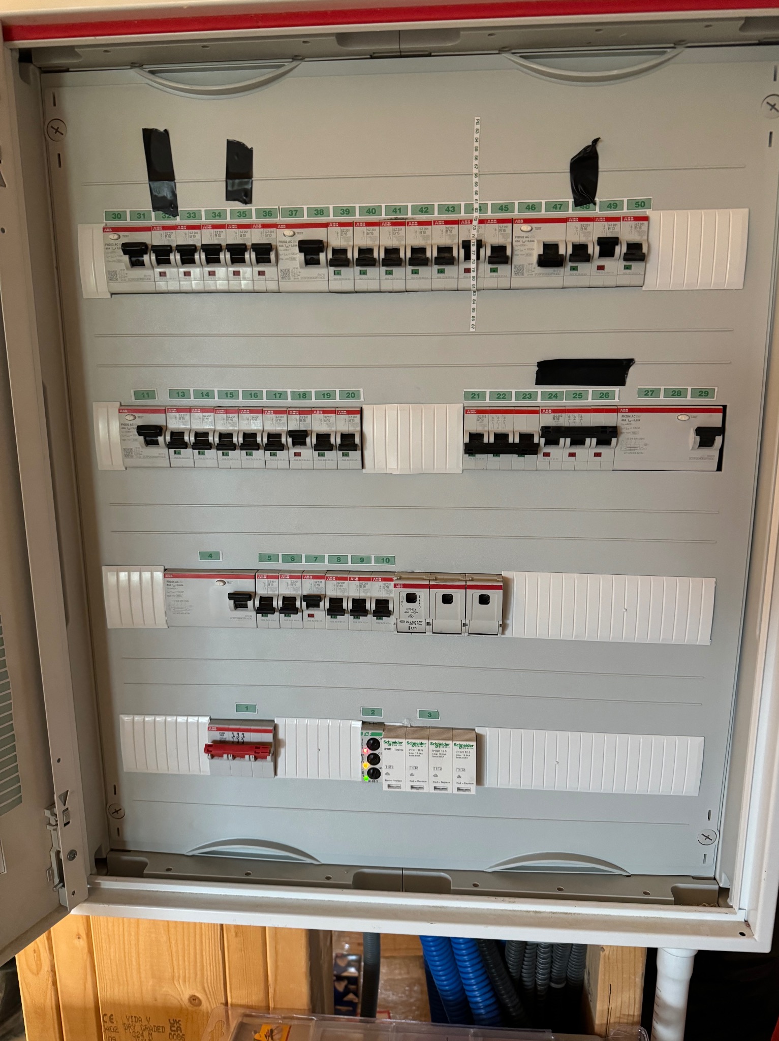 Otwarta skrzynka rozdzielcza z bezpiecznikami, oznaczone numery od 1 do 50, niektóre zabezpieczone czarną taśmą, widoczne różne typy wyłączników, na dole fragment drewnianej ściany i niebieskiego...
