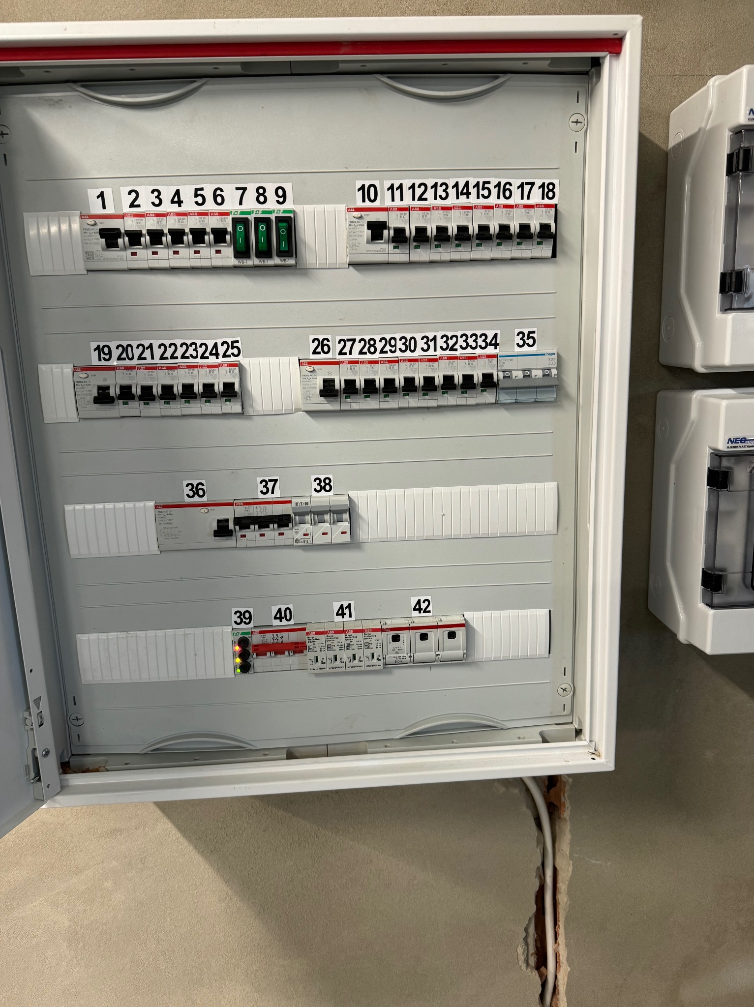 Otwarta skrzynka rozdzielcza z zainstalowanymi bezpiecznikami i wyłącznikami oznaczonymi numerami, widoczne również przewody wychodzące ze ściany.