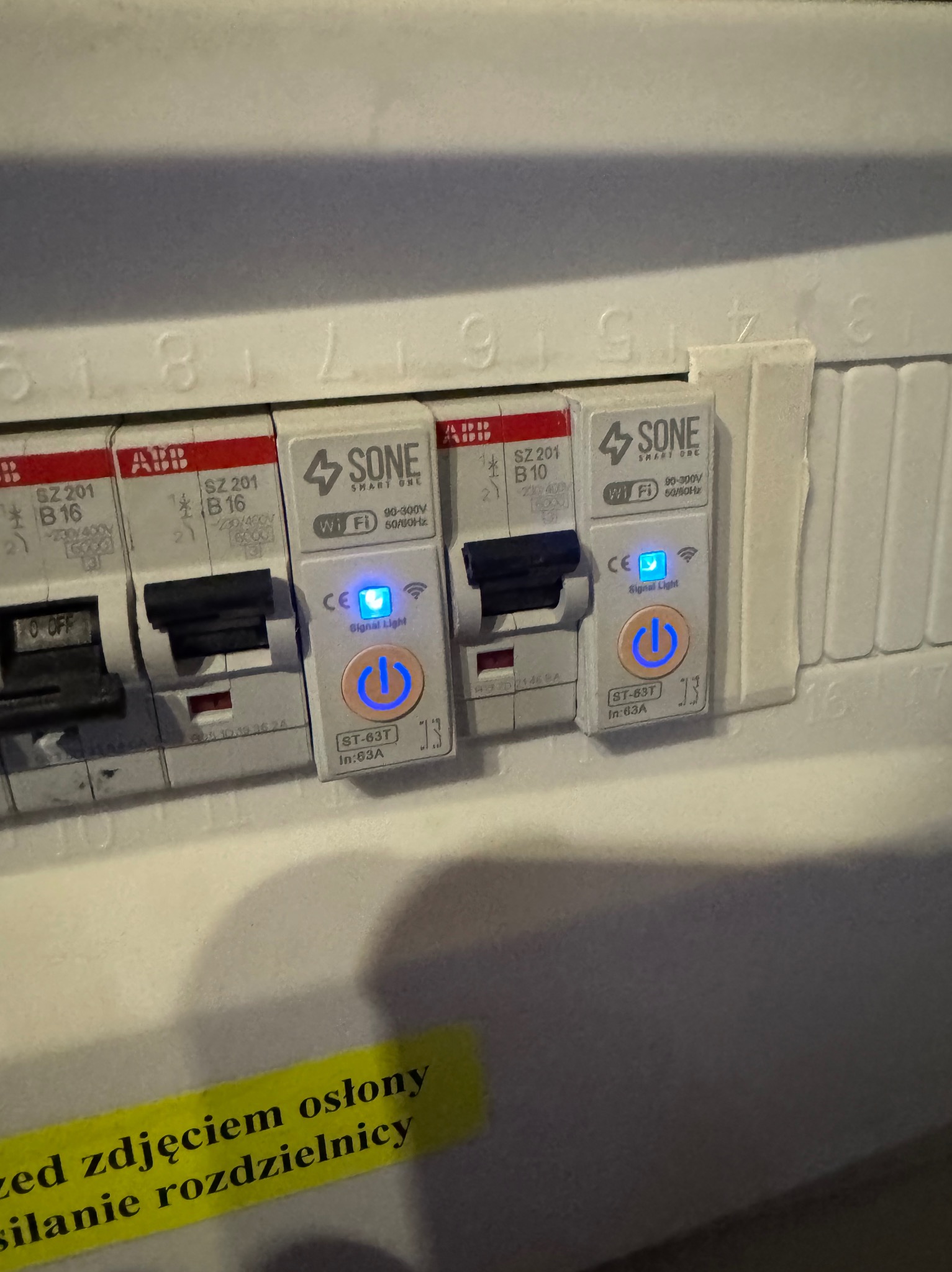Szczegółowe ujęcie tablicy rozdzielczej z zamontowanymi modułowymi wyłącznikami nadprądowymi SONE Smart One z włączonymi diodami LED, widoczna żółta naklejka z napisem.