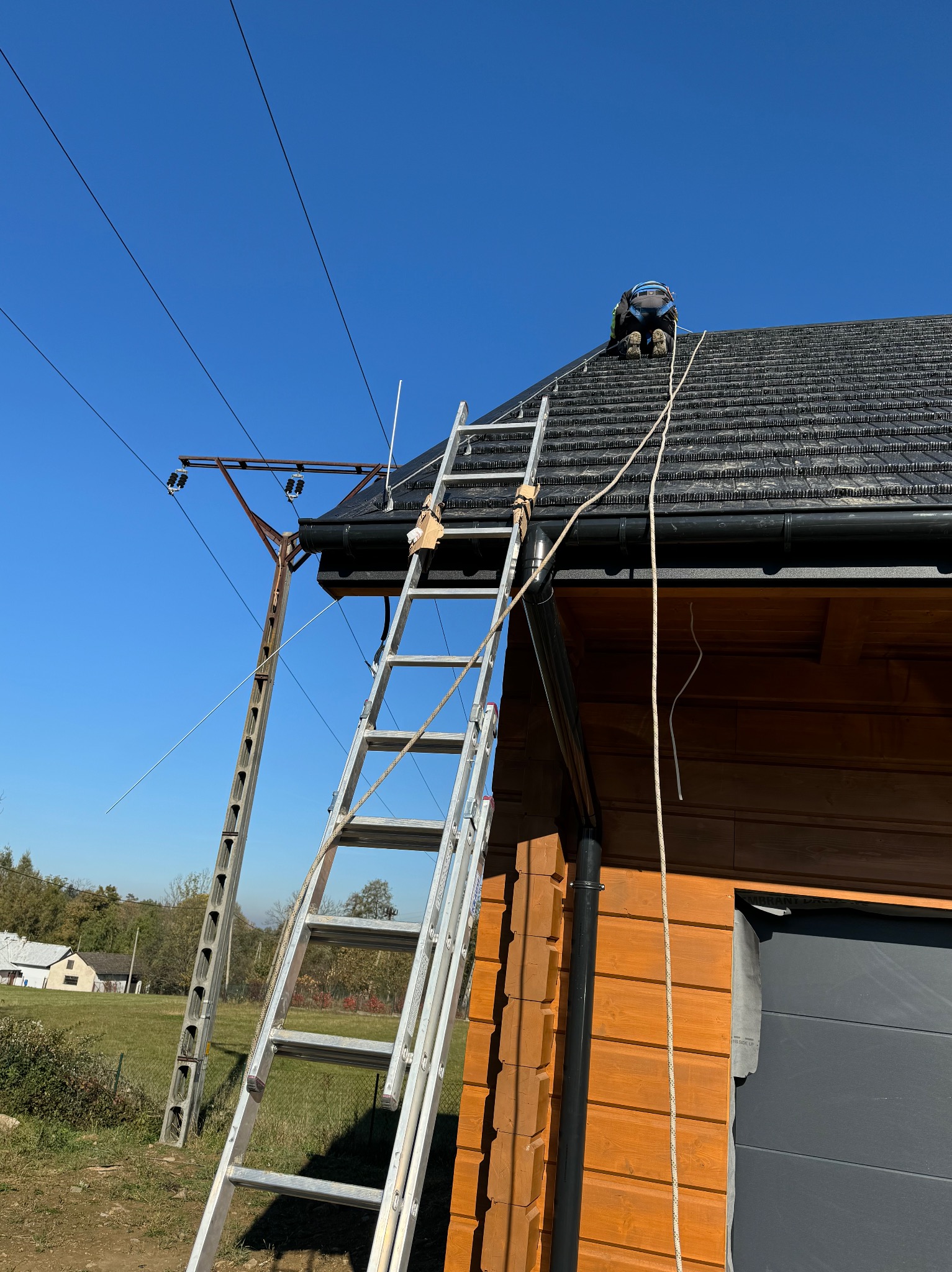 Pracownik w kasku klęczy na dachu drewnianego domu, obok drabina oparta o rynnę, widoczny słup energetyczny i niebieskie niebo.