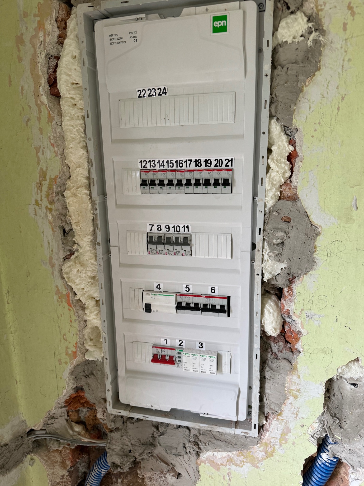 Biała skrzynka rozdzielcza z bezpiecznikami zamontowana w ścianie z widocznymi ubytkami i pianką montażową, fragment niebieskiej rury karbowanej w dolnym rogu.