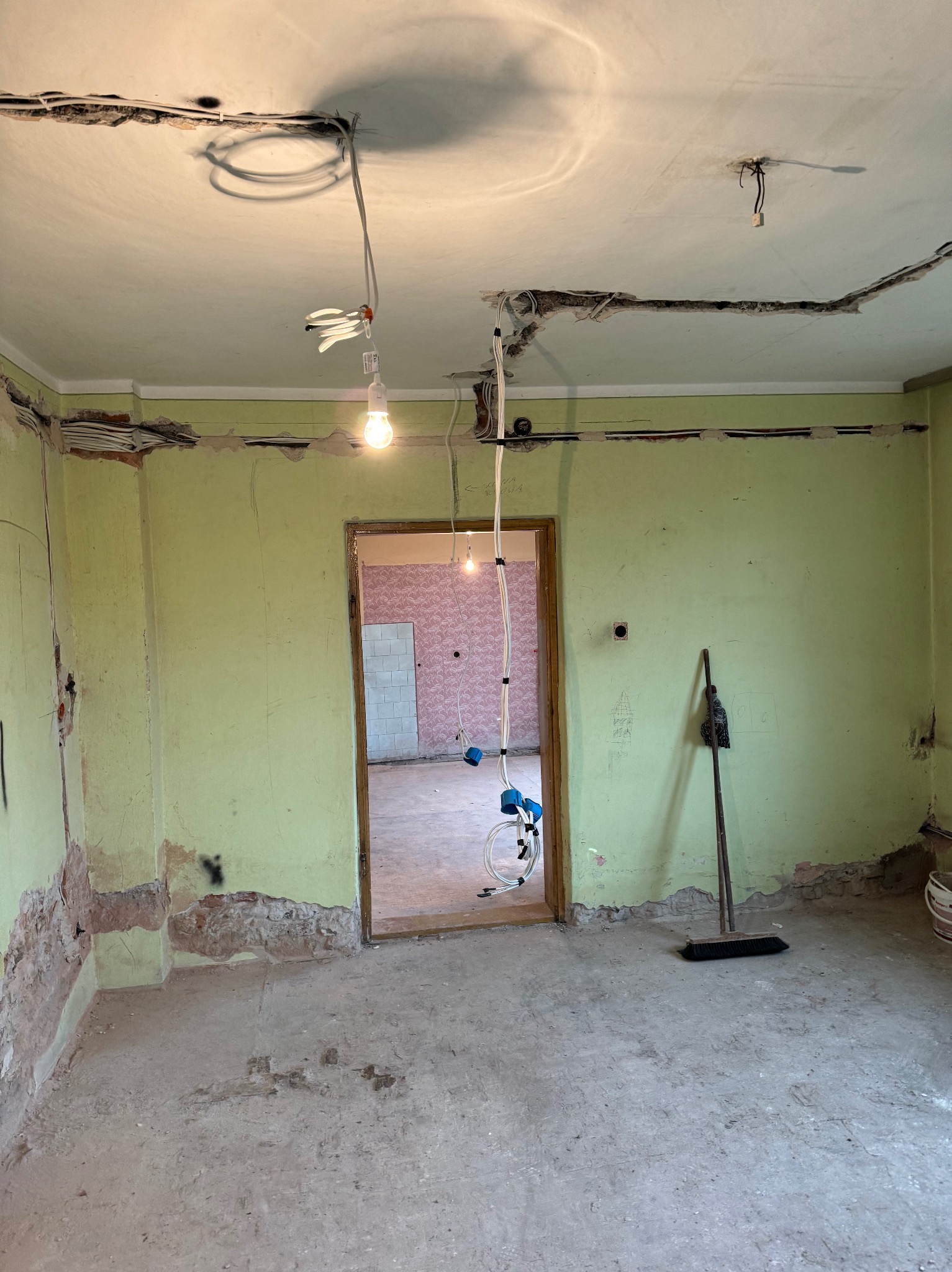 Remont pokoju z odkrytymi przewodami elektrycznymi w listwach na suficie i ścianach, wisząca żarówka na kablu, widoczna instalacja elektryczna w sąsiednim pokoju, ściany w kolorze pistacjowym...