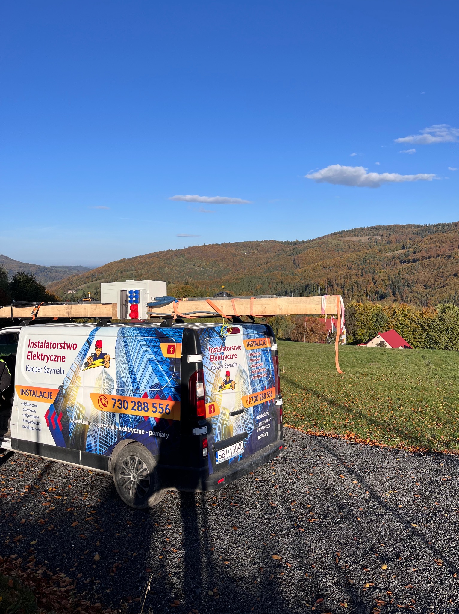 Samochód firmy instalatorskiej z logo i numerem telefonu na tle górskiego krajobrazu, na dachu przewożony element konstrukcyjny zabezpieczony pasami.