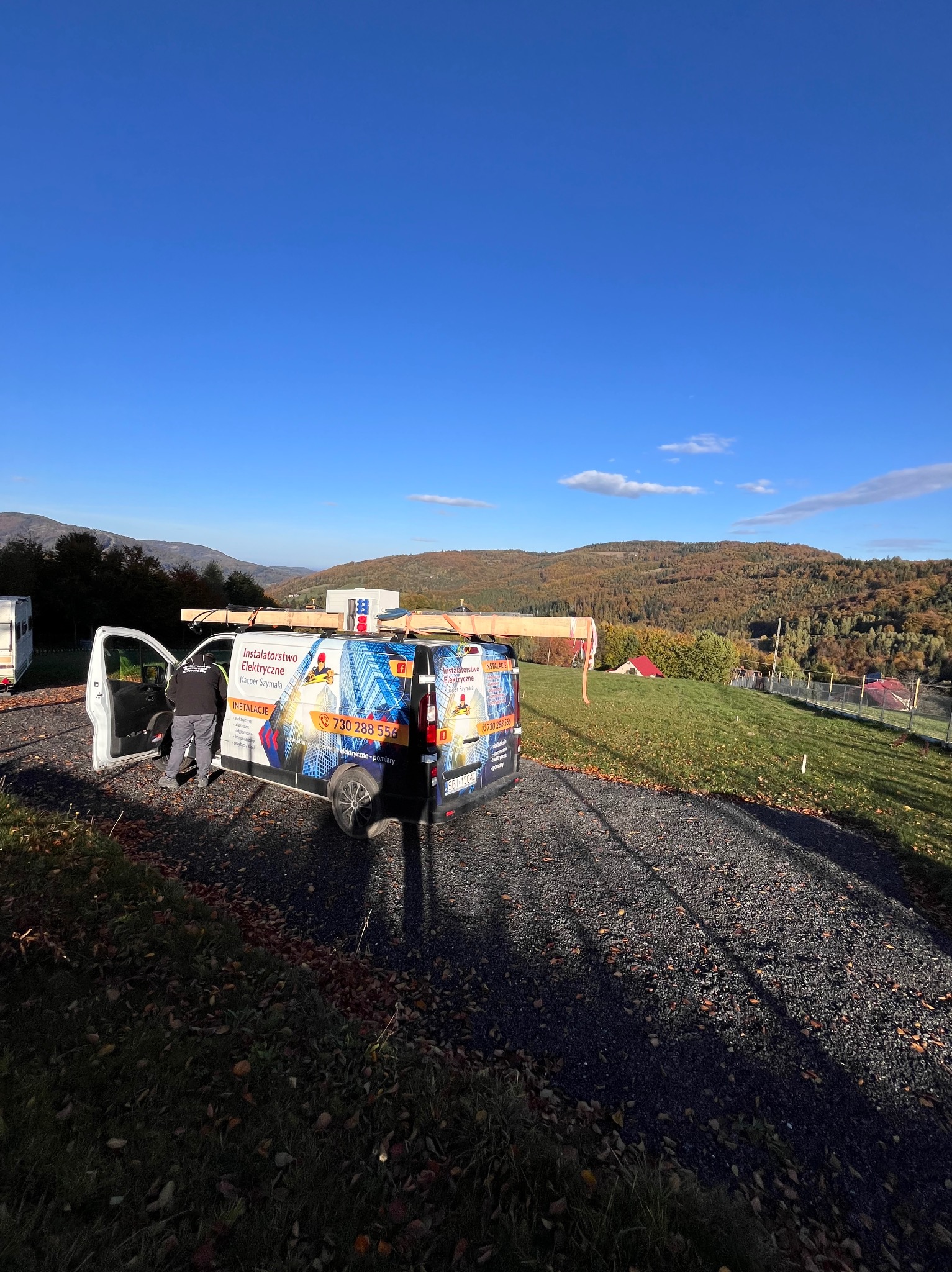 Samochód firmowy z logo i numerem telefonu na tle górskiego krajobrazu, z drewnianymi elementami na dachu, podczas dnia.