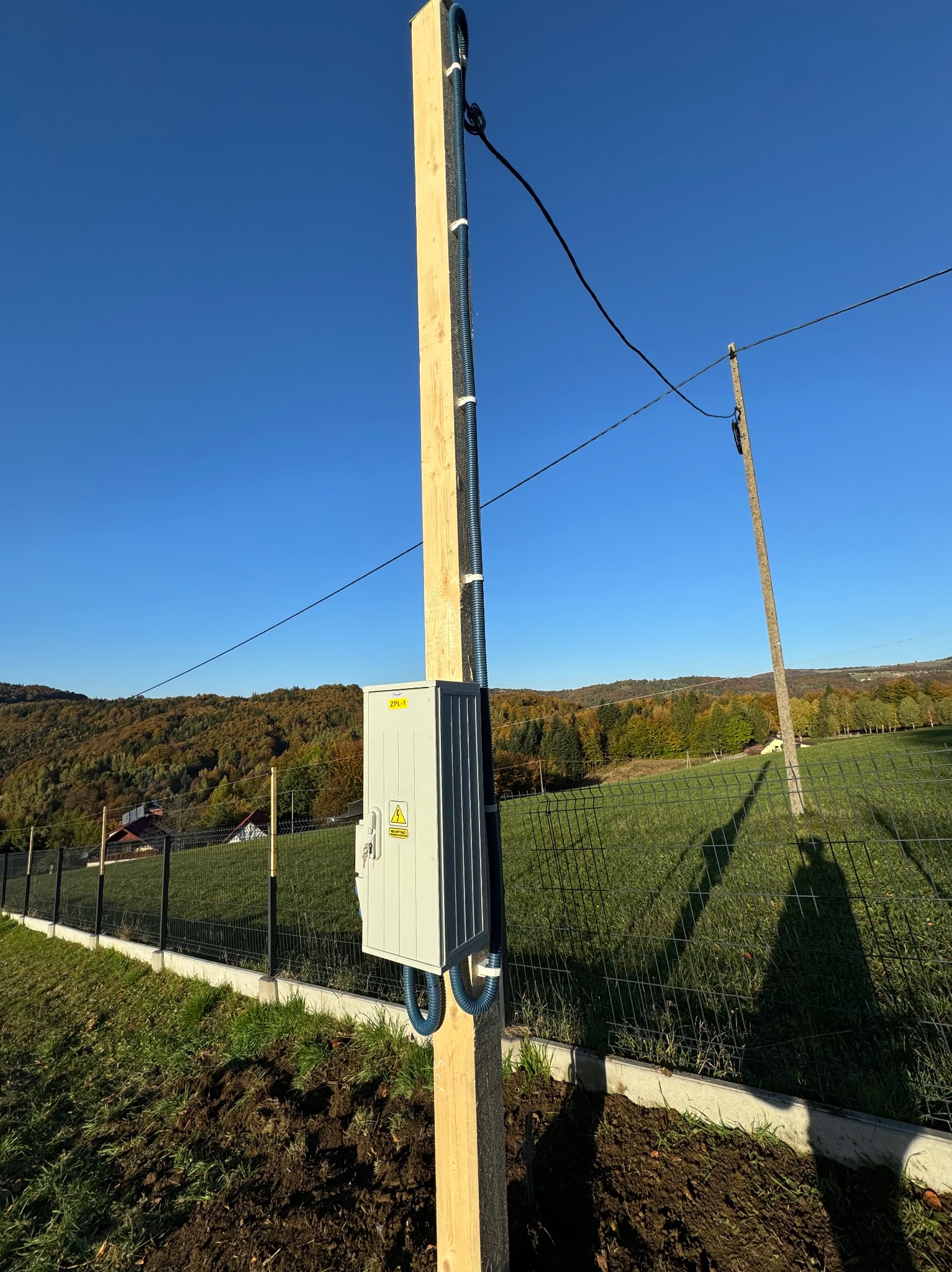 Nowo zainstalowana skrzynka elektryczna na drewnianym słupie, z niebieskimi rurami karbowanymi, na tle krajobrazu z lasem i niebieskim niebem.