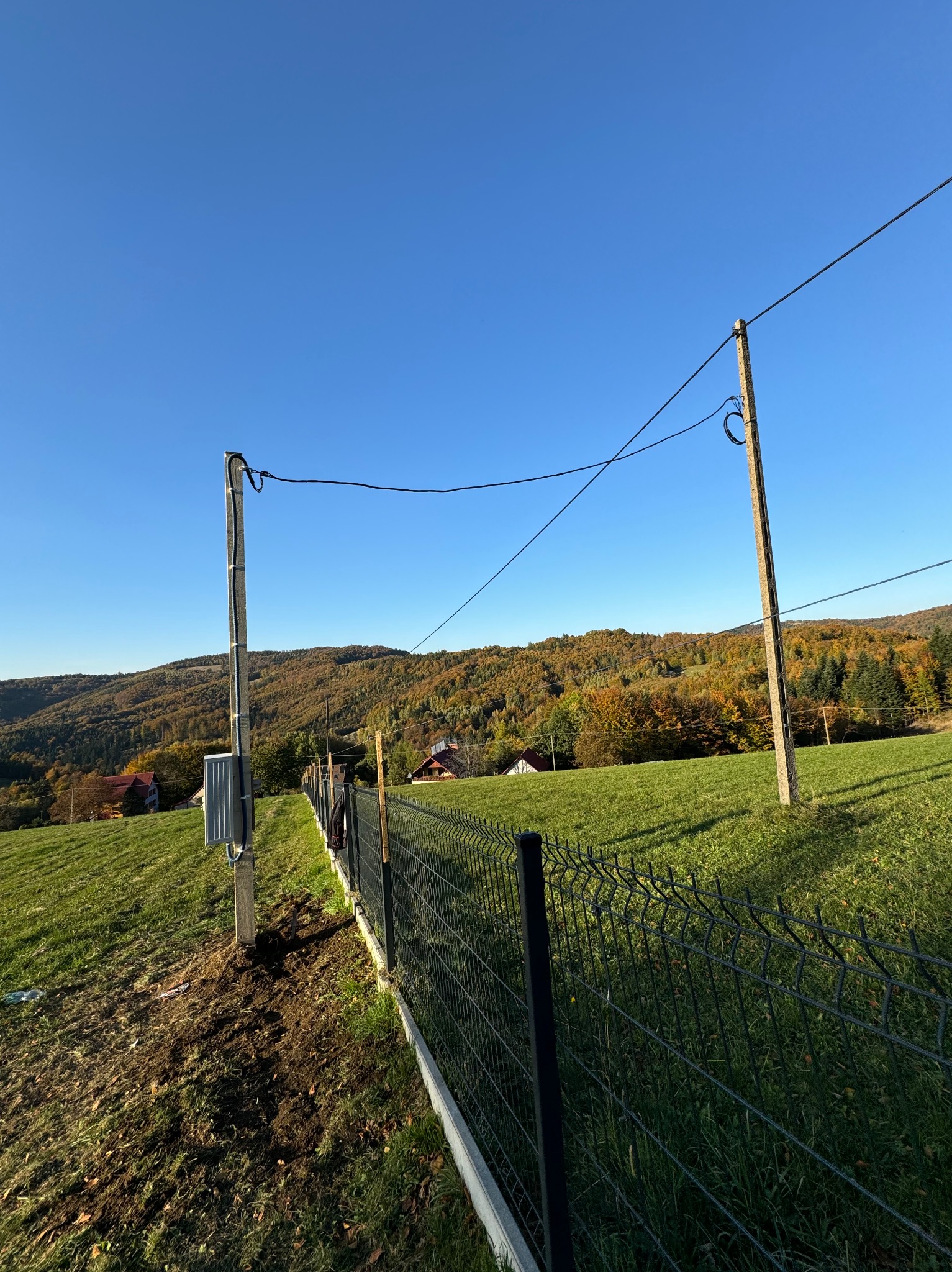 Słup energetyczny z przyłączem kablowym i skrzynką licznikową na tle krajobrazu z ogrodzeniem panelowym.
