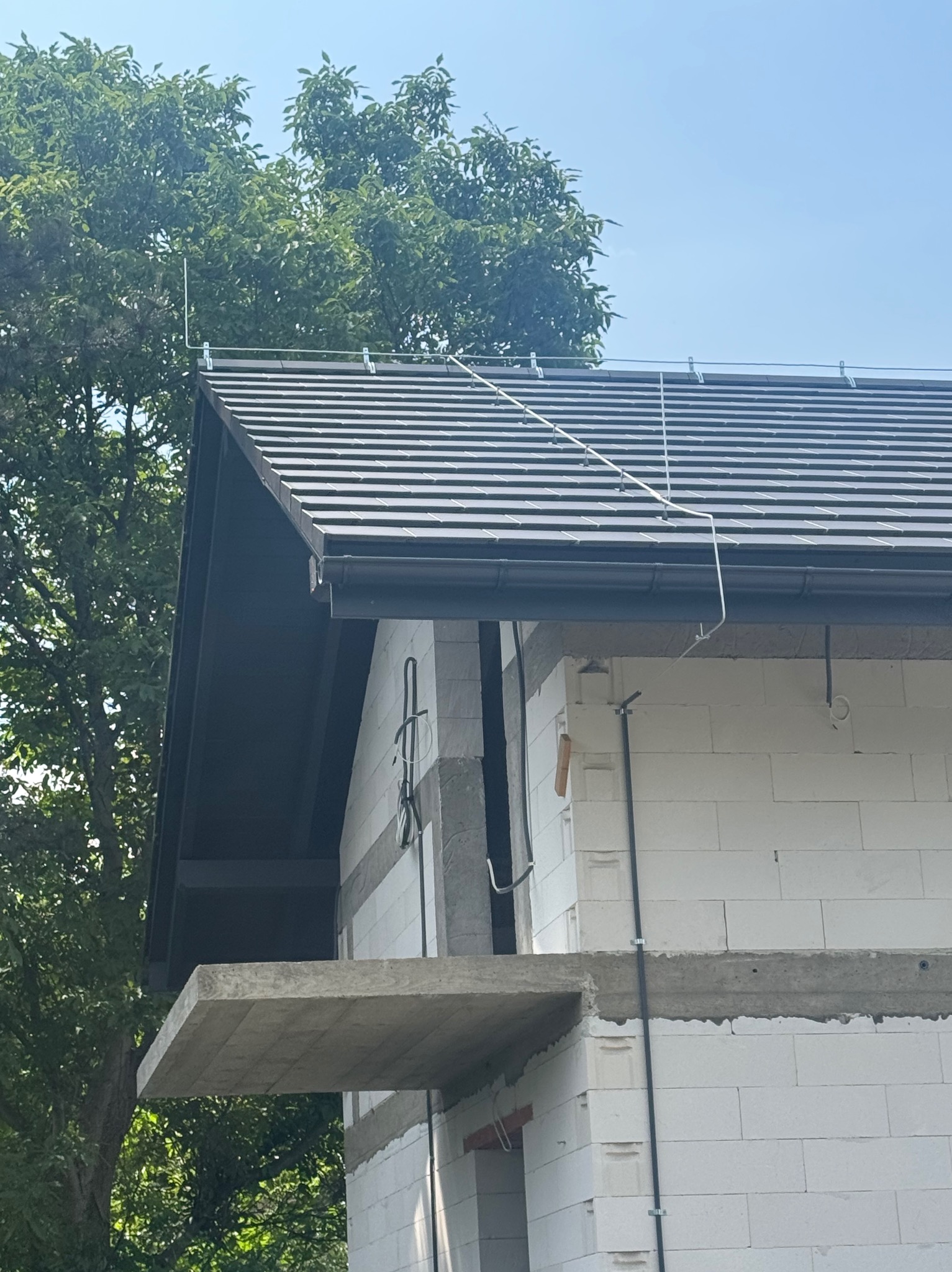 Dom w trakcie budowy z zamontowaną instalacją odgromową na dachu, widoczne przewody i uziomy. Budynek z białego bloczka, w tle drzewo i niebo.