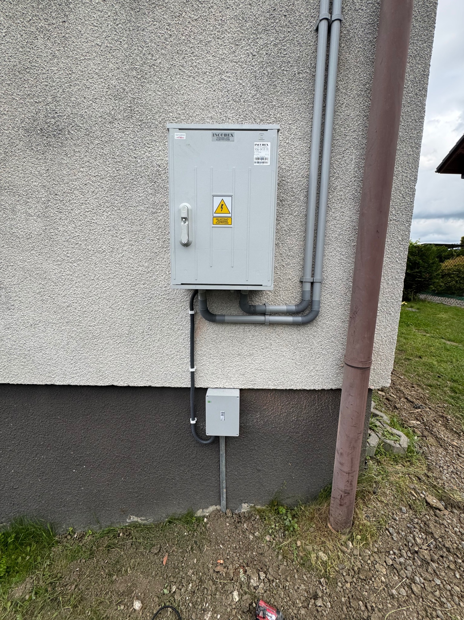Zewnętrzna skrzynka elektryczna INCOBEX na elewacji domu, z widocznymi rurami osłonowymi i uziemieniem, zabezpieczona przed warunkami atmosferycznymi.