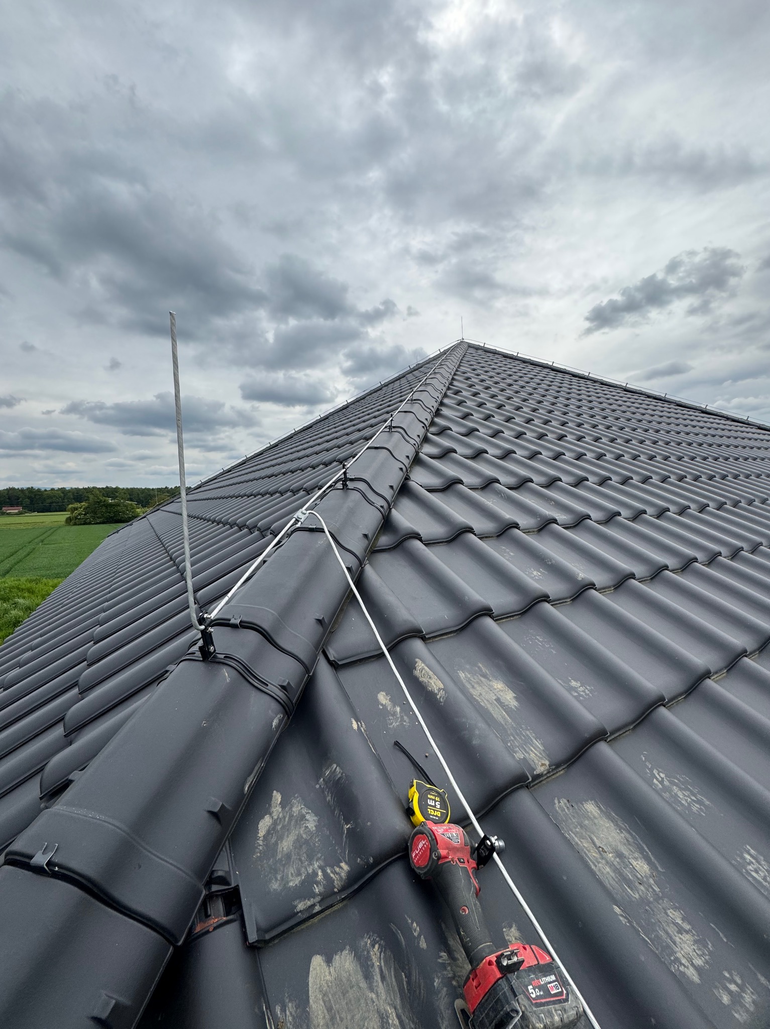 Montaż instalacji odgromowej na dachu z czarnej dachówki, widoczne narzędzia i przewody, pochmurne niebo w tle. Praca w toku, zbliżenie na mocowania.