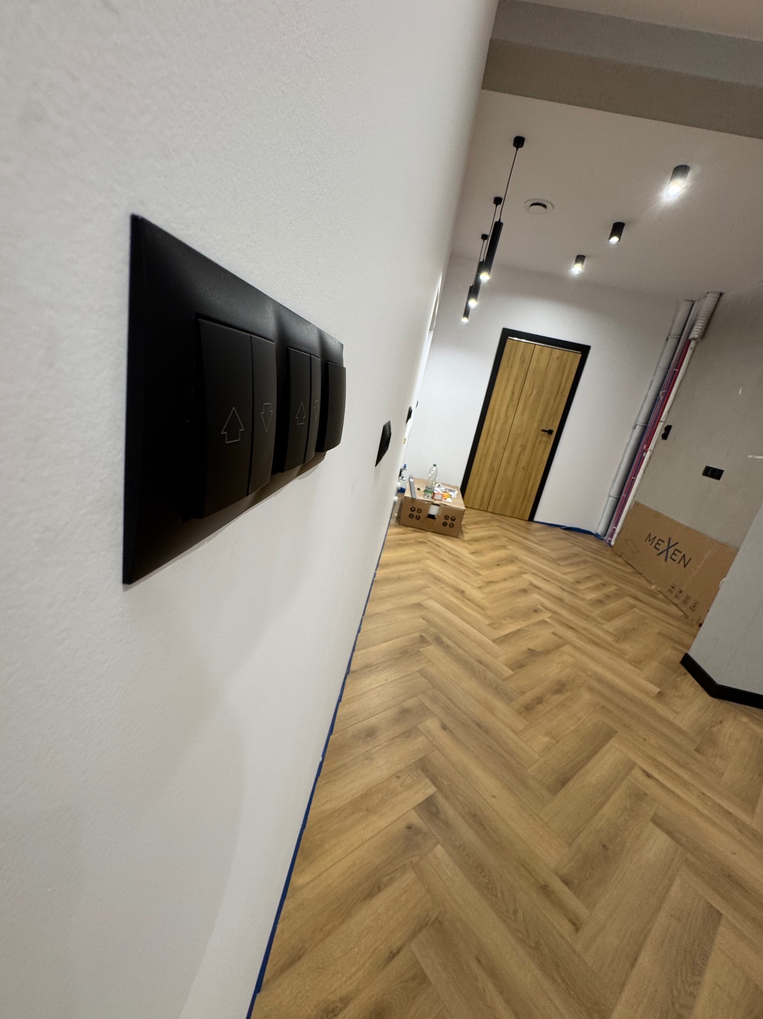 Czarny panel z włącznikami światła na białej ścianie, z widocznym parkietem i lampami sufitowymi w tle, w nowoczesnym wnętrzu.