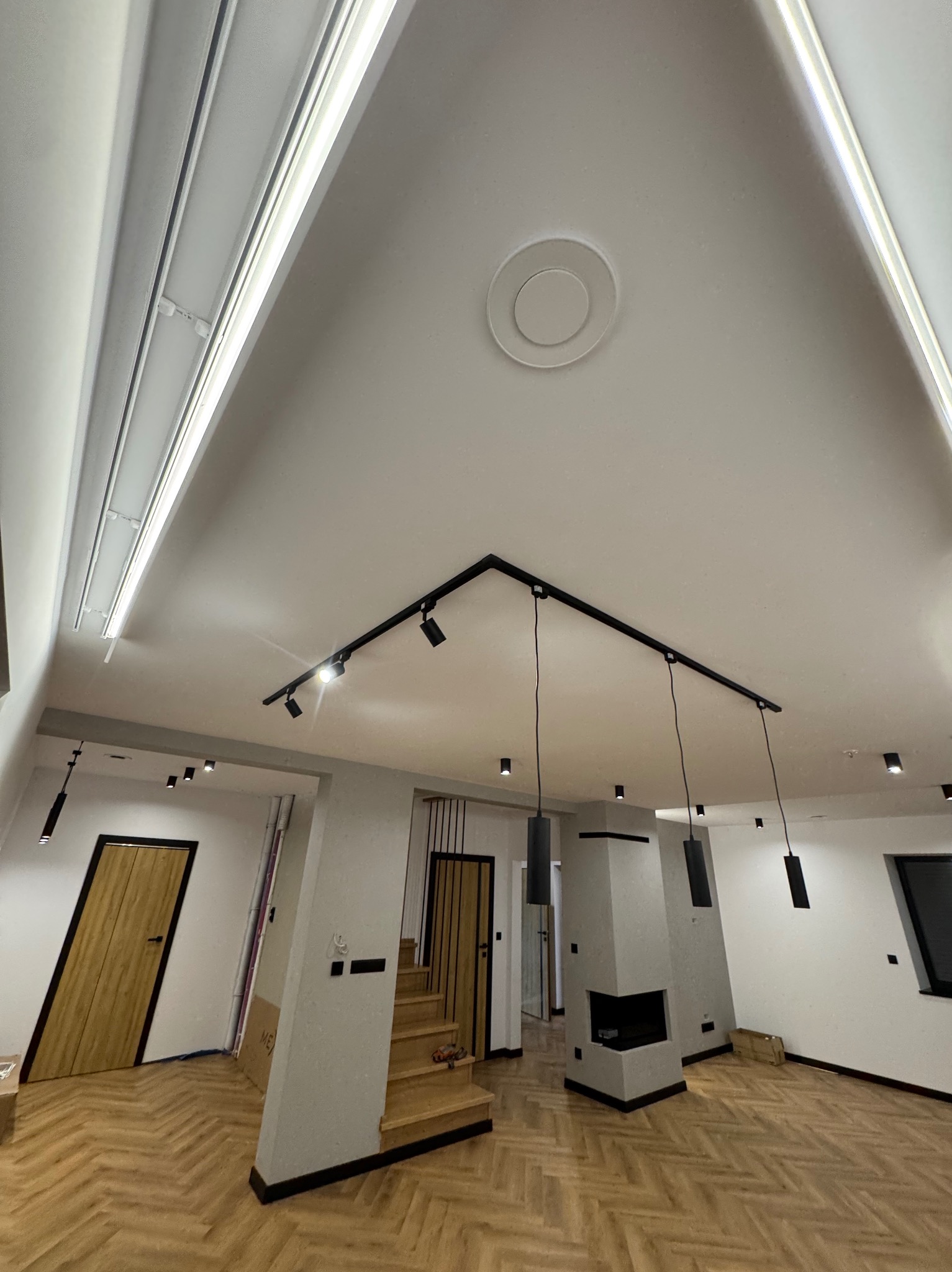 Nowoczesne oświetlenie sufitowe w salonie z parkietem w jodełkę i kominkiem. Widoczne halogeny, listwy LED i czarne lampy wiszące.