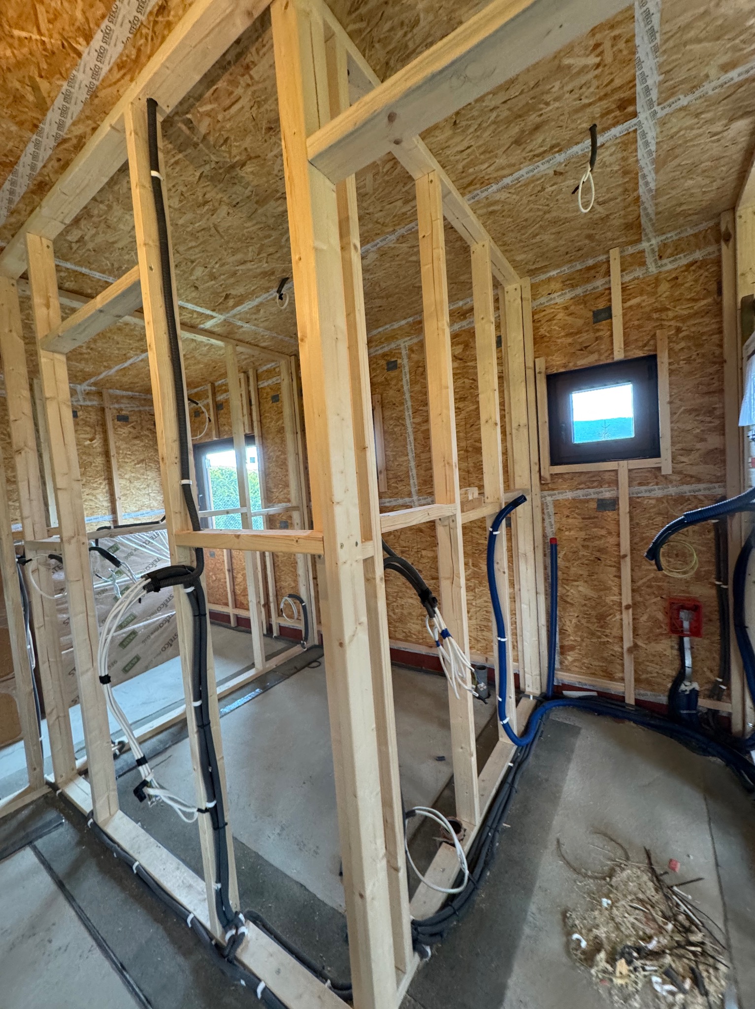Wnętrze w trakcie budowy z widocznymi drewnianymi konstrukcjami ścian i stropu. Ułożone przewody elektryczne i instalacyjne w peszlach. Małe okno w ścianie.