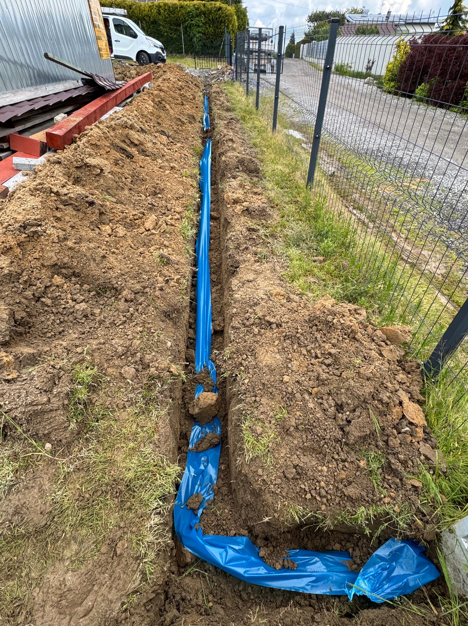 Wykop z niebieską folią ochronną w ziemi, prawdopodobnie pod instalację. Widoczny fragment ogrodzenia i trawa. Wykop świeżo zrobiony, ziemia wokół.