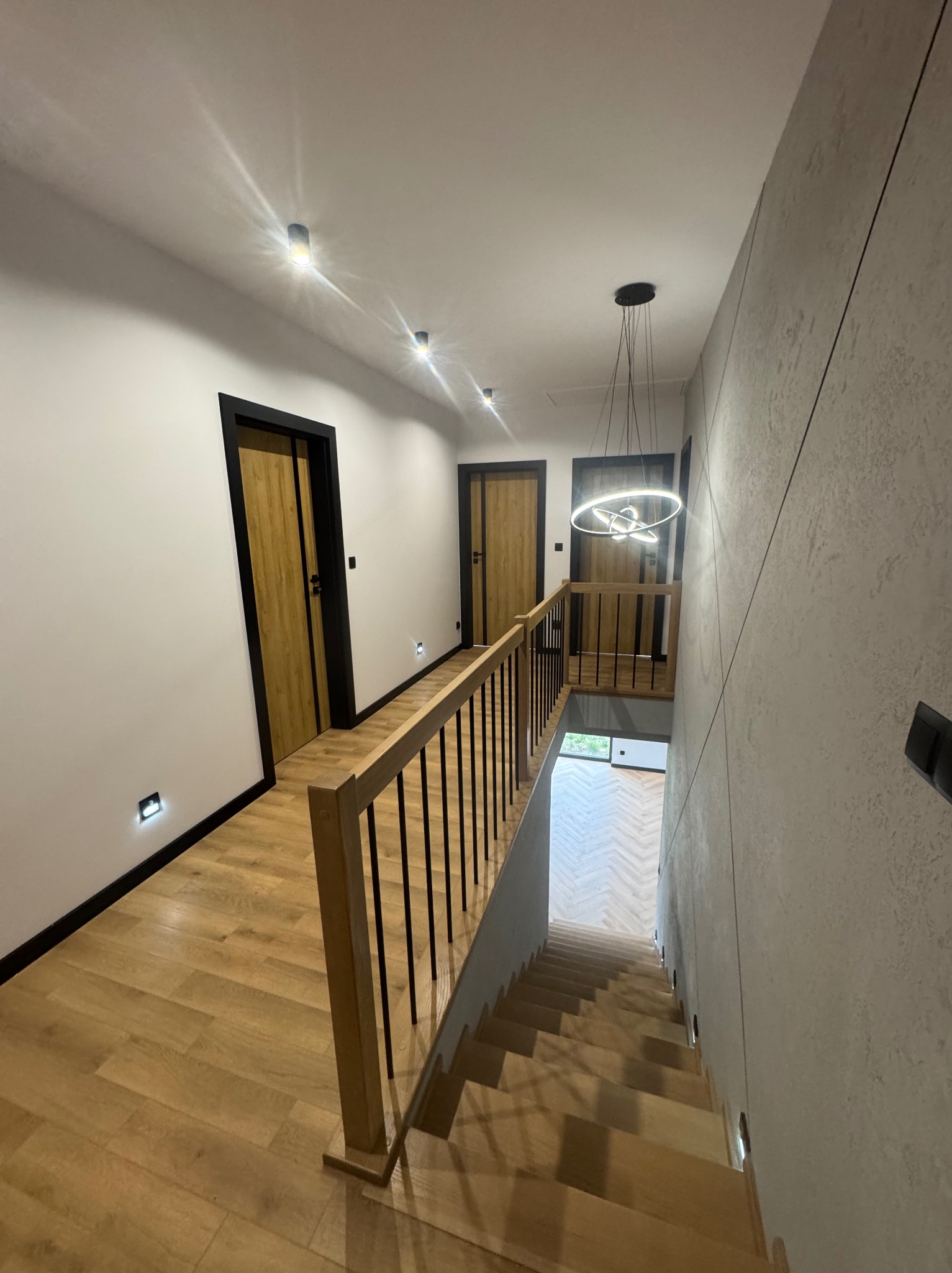 Nowoczesne oświetlenie LED w korytarzu z drewnianymi schodami i podłogą. Widoczne lampy sufitowe i ścienne, oraz designerska lampa wisząca nad schodami.