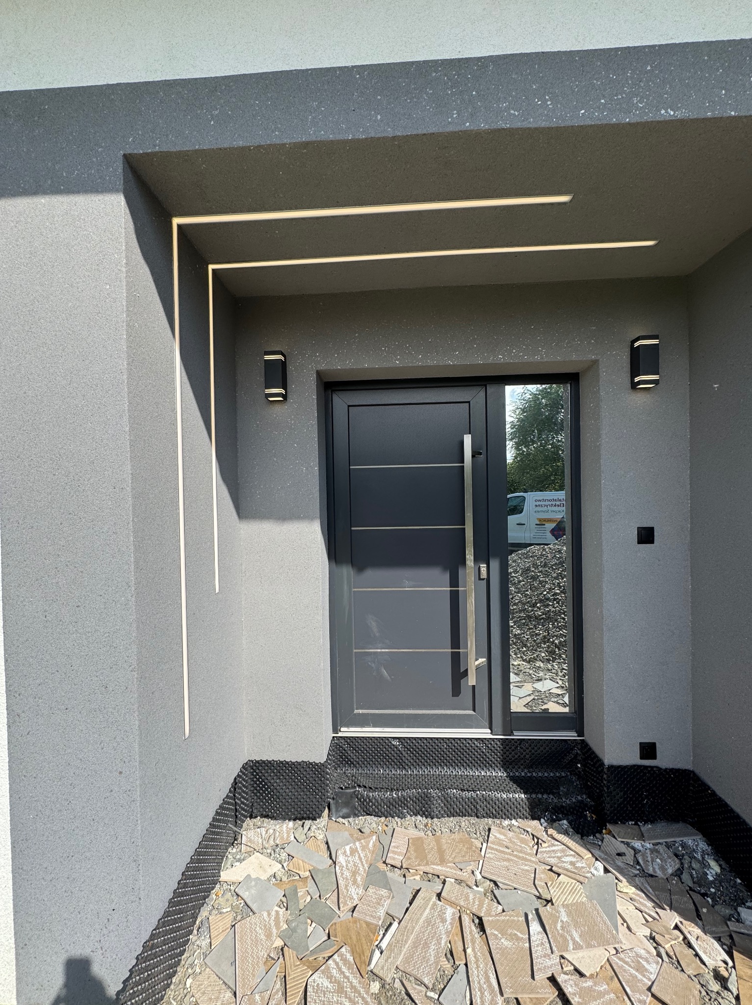 Eleganckie wejście do domu z nowoczesnym oświetleniem zewnętrznym: lampy ścienne i ledowe linie wzdłuż framugi. Fragment gruzu po pracach wykończeniowych.