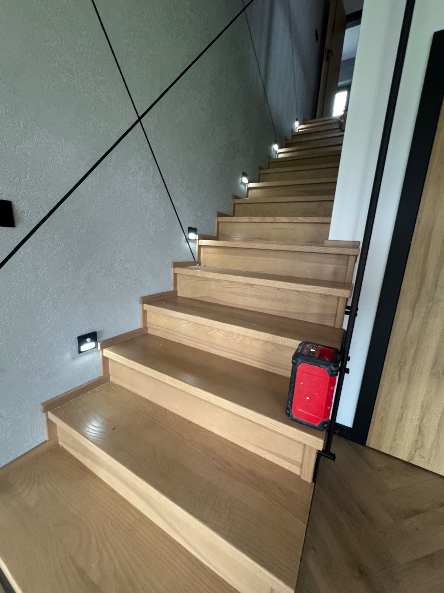 Drewniane schody z oświetleniem punktowym na ścianie z dekoracyjną, czarną linią. Widoczny fragment drzwi i poziomica laserowa.