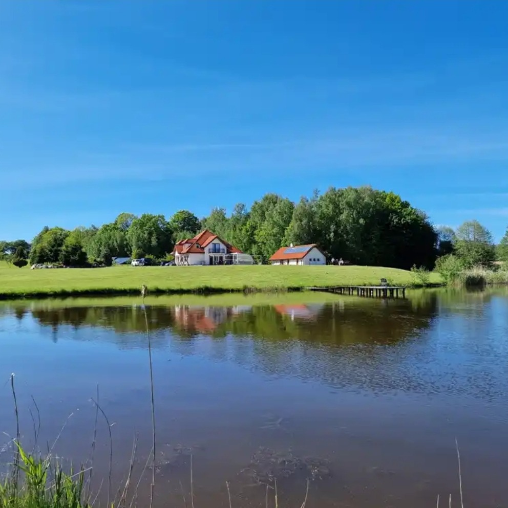 Malowniczy krajobraz z jeziorem odbijającym dom z czerwoną dachówką i zielone drzewa pod błękitnym niebem.