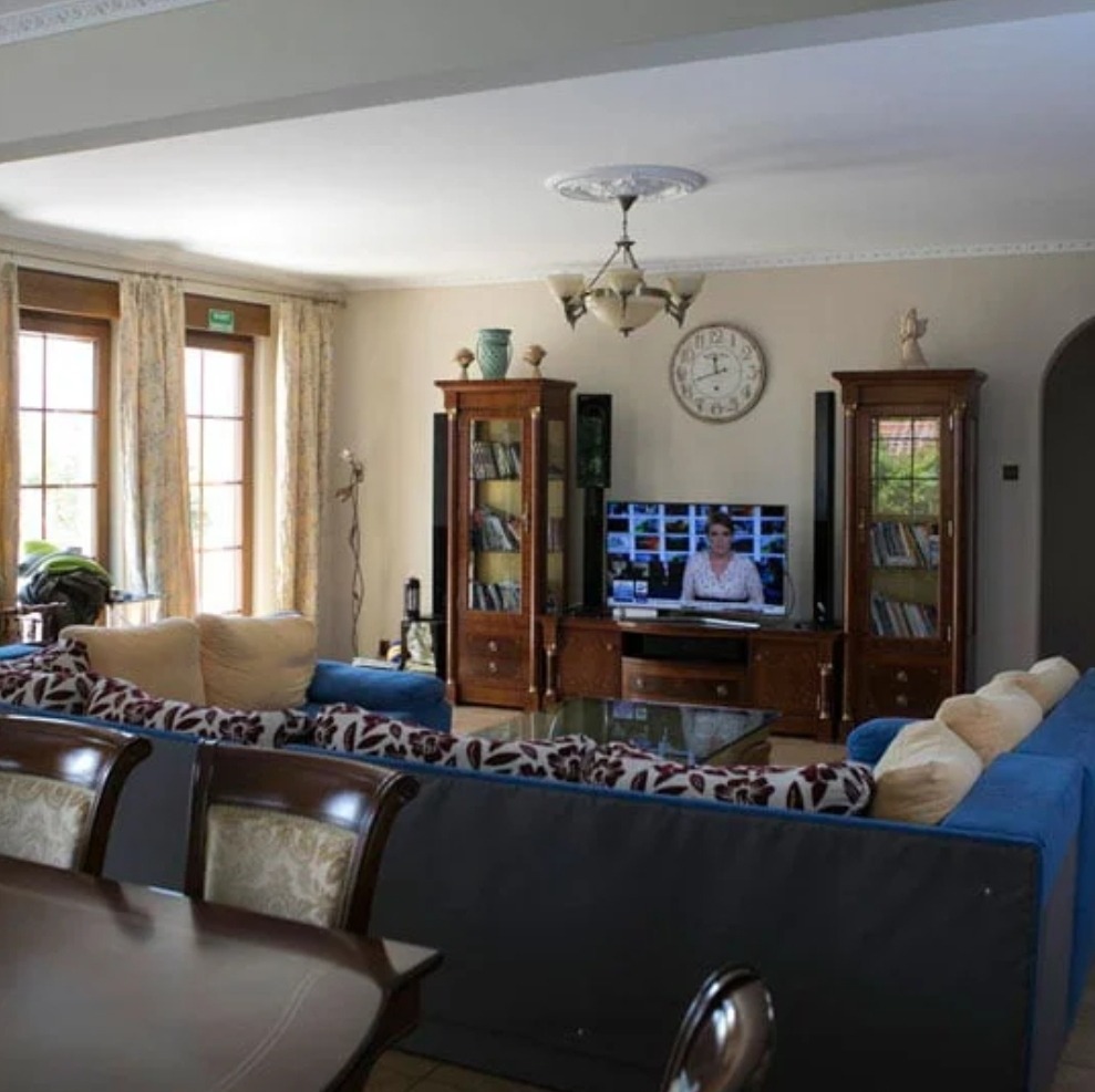 Jasny salon z niebieską sofą, drewnianymi witrynami i telewizorem wyświetlającym kobietę, zegar na ścianie wskazuje godzinę 12:53.