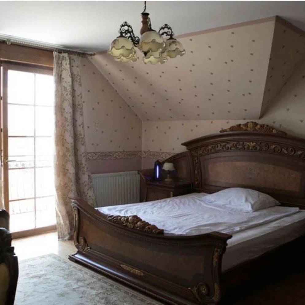 Pokój z dużym, drewnianym łóżkiem w stylu retro, wzorzystą tapetą i ozdobnym żyrandolem, okno z widokiem na balkon.