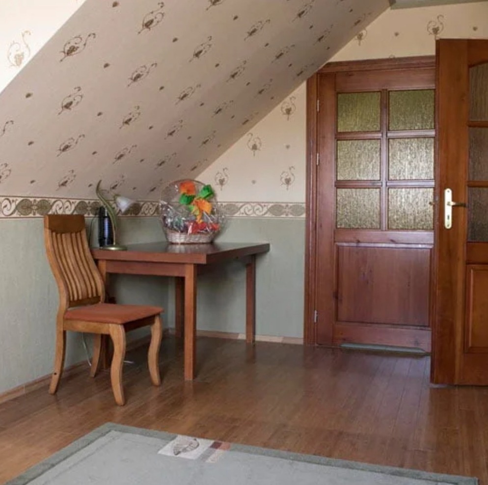 Pokój z drewnianym stołem, krzesłem i koszem prezentowym w Hucie Żuławskiej, w tle skośny sufit i drzwi z szybkami.