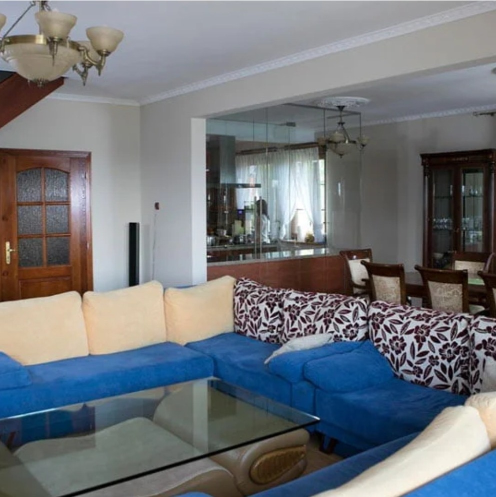 Przestronny salon z narożną, niebieską sofą, szklanym stolikiem kawowym i widokiem na jadalnię przez szklaną ścianę.