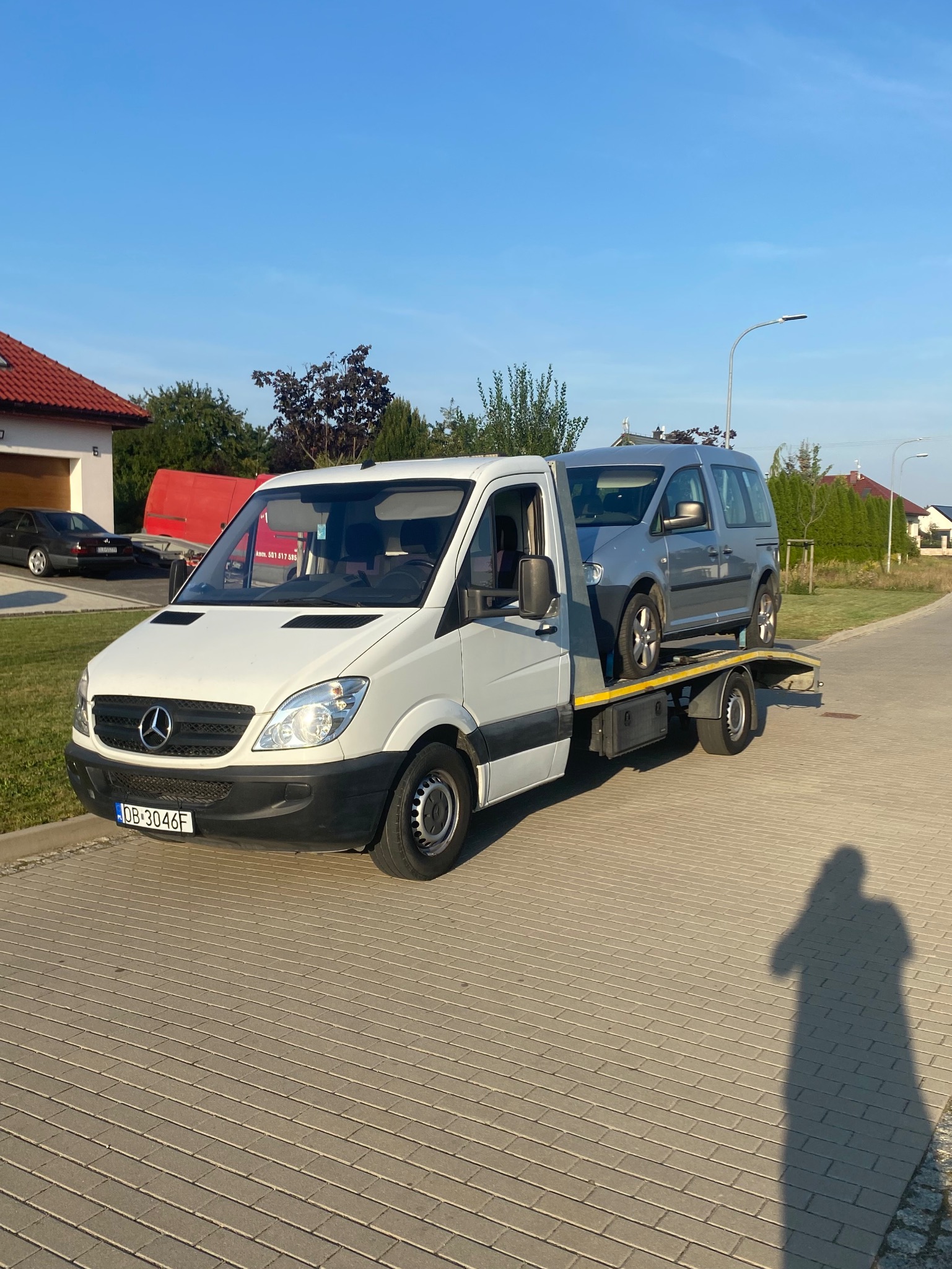 Biała laweta Mercedes-Benz z szarym samochodem osobowym na platformie, zaparkowana na brukowanej drodze w słoneczny dzień.