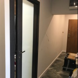 TOM-BUD - Częściowo przesuwne drzwi wewnętrzne z czarną ramą i matową szybą, widoczne w korytarzu z marmurową podłogą i sprzętem AGD na ścianie.