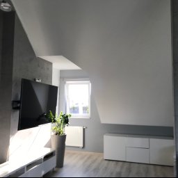 TOM-BUD - Nowoczesny salon na poddaszu z szarą ścianą imitującą beton, białymi meblami, telewizorem i roślinami doniczkowymi w promieniach słońca.
