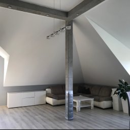 TOM-BUD - Jasne, przestronne poddasze po remoncie, z szarym narożnikiem, białymi meblami i wykończonymi ścianami, oświetlone nowoczesną lampą wiszącą.