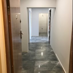 TOM-BUD - Korytarz po remoncie z szarymi płytkami imitującymi marmur na podłodze, świeżo pomalowanymi ścianami na biało i widocznym wejściem do łazienki w trakcie prac wykończeniowych.