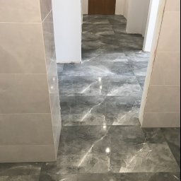 TOM-BUD - Ułożona szara marmurkowa podłoga z płytek w korytarzu, widoczne jasne płytki na ścianie i fragment drewnianych drzwi w tle.