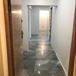 TOM-BUD - Korytarz po remoncie z szarymi płytkami imitującymi marmur na podłodze, świeżo pomalowane ściany na szaro, widok przez otwarte drzwi wejściowe na łazienkę w głębi.