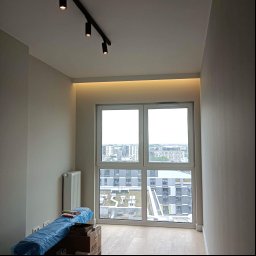 TOM-BUD - Wnętrze pokoju z dużym oknem z widokiem na miasto, świeżo pomalowane ściany w odcieniach szarości, drewnopodobna podłoga, grzejnik, oświetlenie szynowe na suficie i ukryte oświetlenie LED nad oknem...