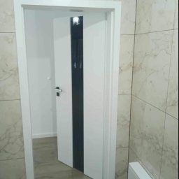 TOM-BUD - Białe, częściowo otwarte drzwi z czarnym pasem, zamontowane w łazience wykończonej marmurowymi płytkami, widok na sąsiednie, pomalowane na biało pomieszczenie.