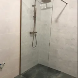 Nowoczesna kabina prysznicowa walk-in z szklaną ścianką i miedzianą armaturą na tle jasnych płytek z geometrycznym wzorem i ciemnej marmurowej podłogi.