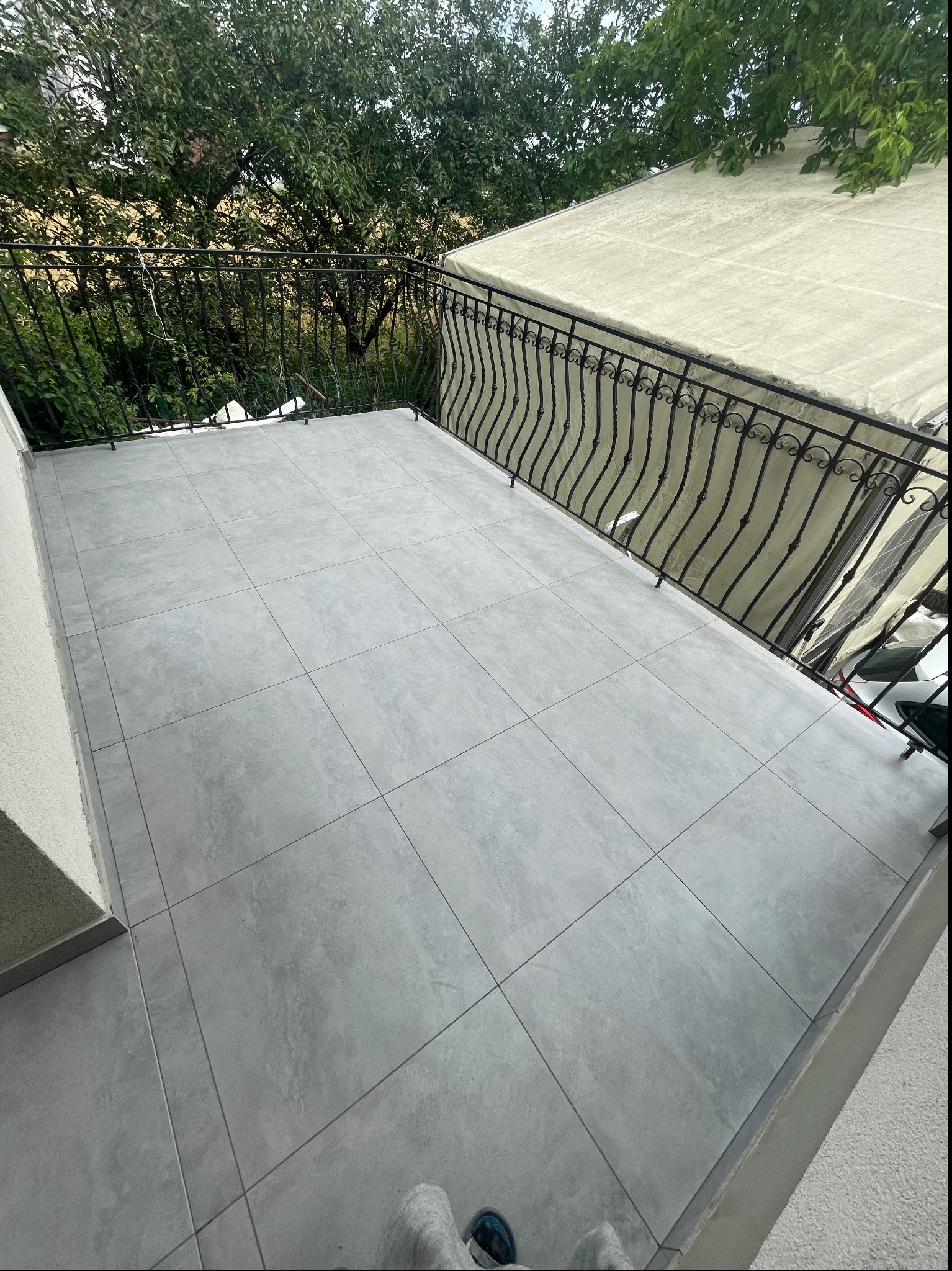 Jasnoszare płytki na balkonie z kutą, ozdobną balustradą, widok na zieleń drzew i zadaszenie