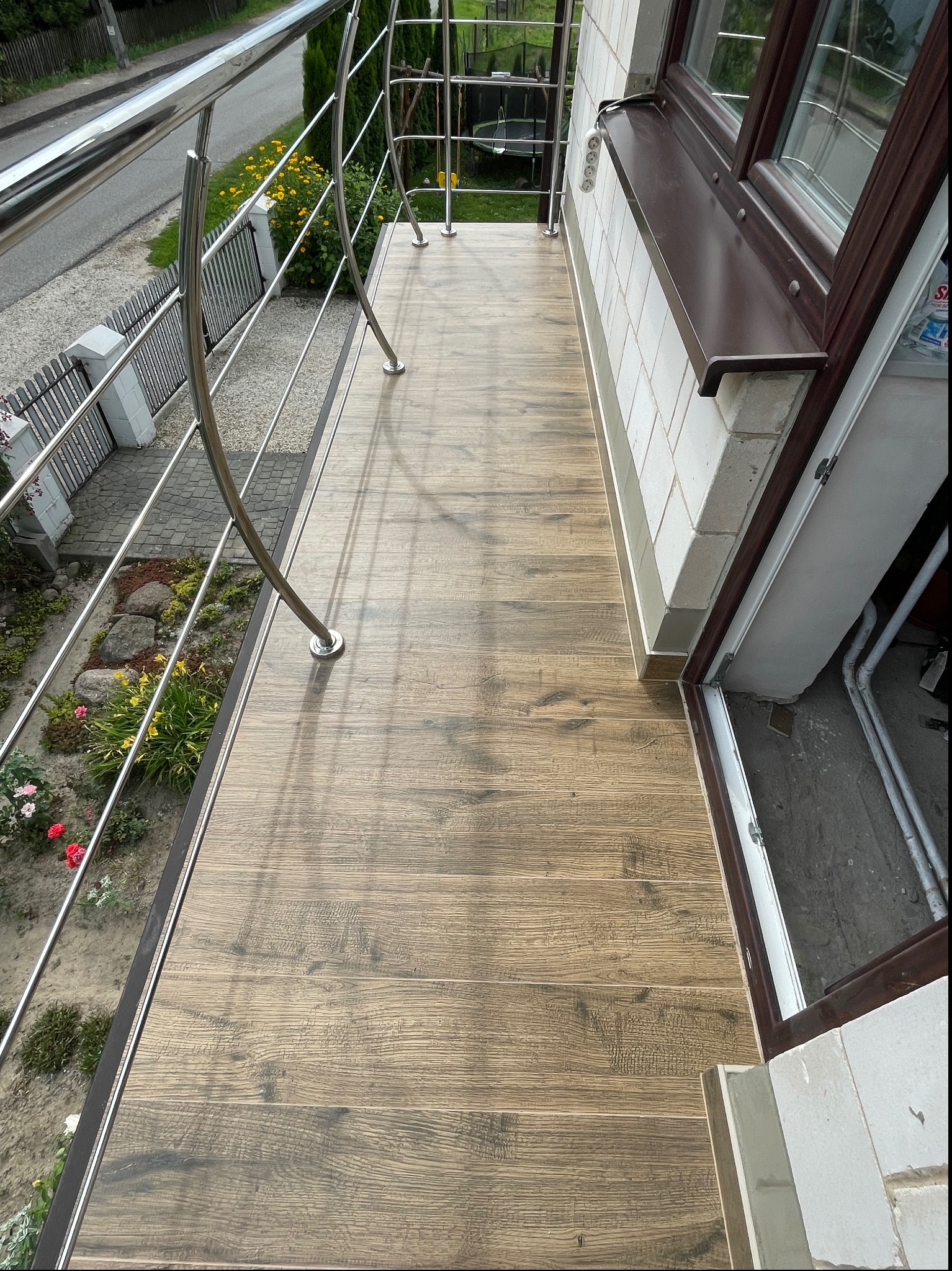Balkon wyłożony płytkami imitującymi drewno, z chromowaną balustradą i widokiem na ogród z kwiatami.