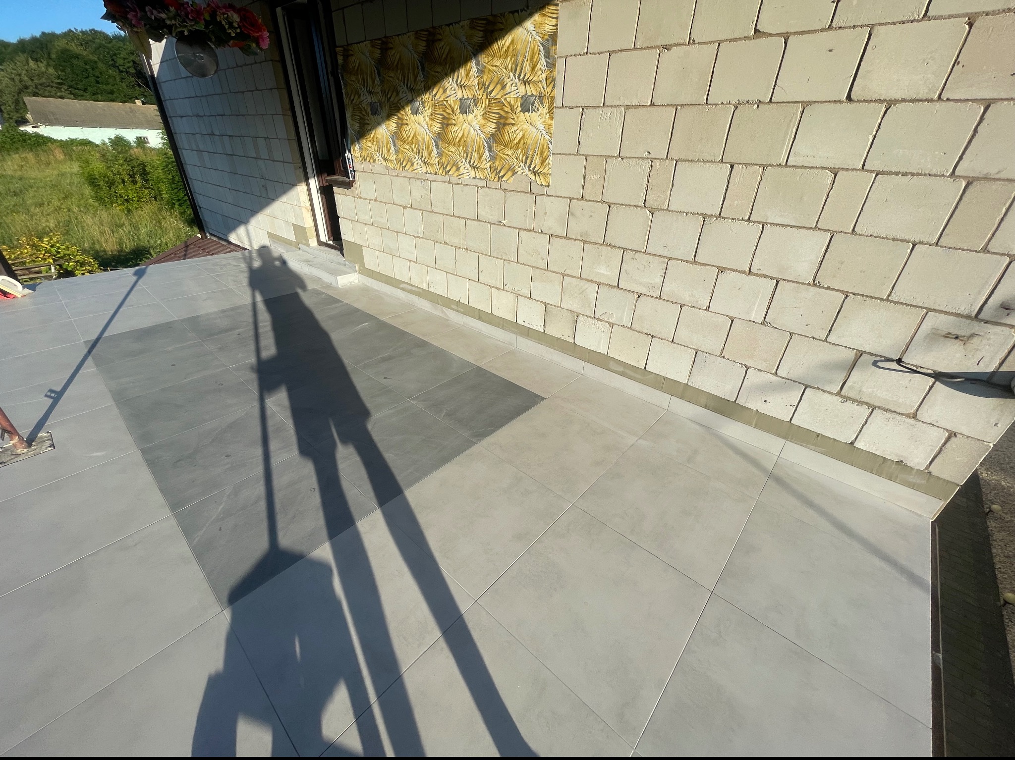 Ułożona szara terakota na tarasie, widoczny cień osoby wykonującej pracę oraz fragment ściany budynku z dekoracyjnym panelem.