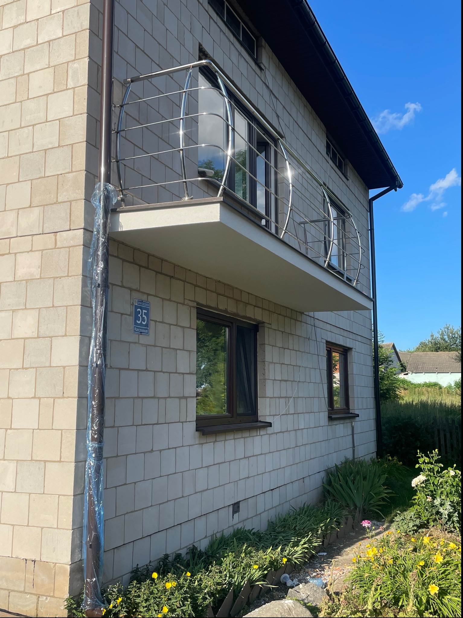 Elewacja budynku z szarej cegły z balkonami z metalową balustradą; rura spustowa zabezpieczona folią ochronną, widoczny numer domu.