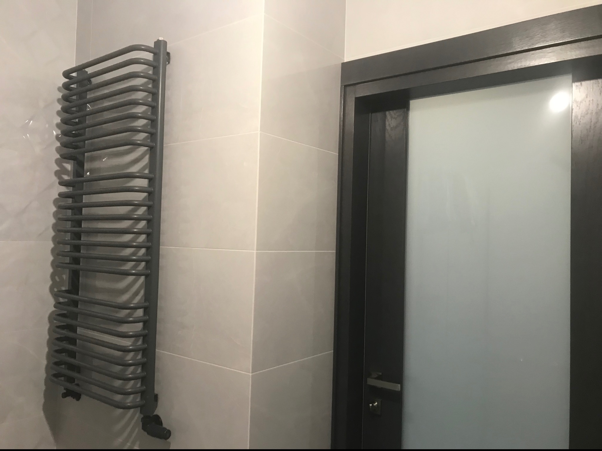 Nowoczesna łazienka z szarymi płytkami, grzejnikiem drabinkowym w kolorze antracytowym i drzwiami z matowego szkła w ciemnej ramie.