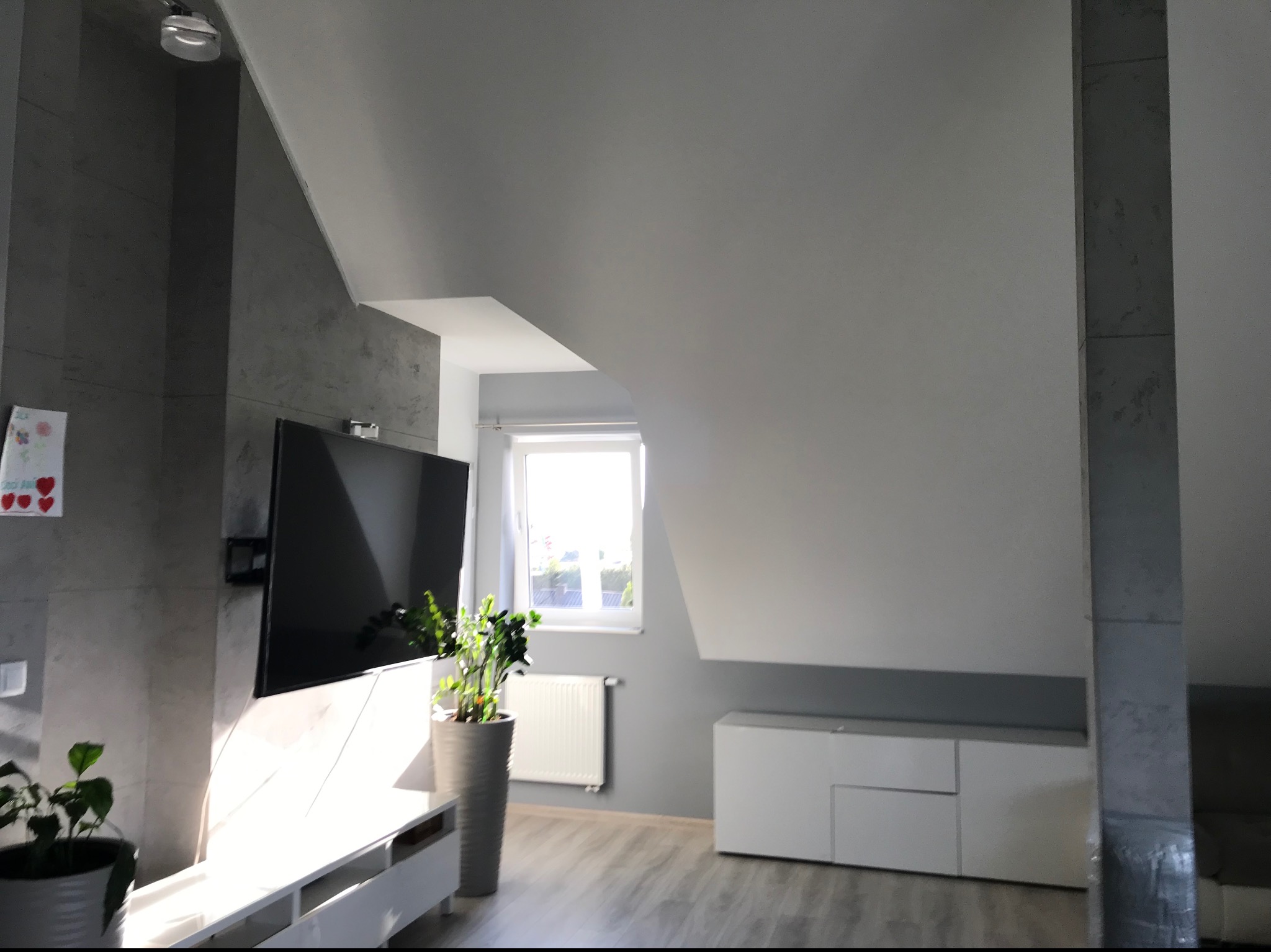 Nowoczesny salon na poddaszu z szarą ścianą imitującą beton, białymi meblami, telewizorem i roślinami doniczkowymi w promieniach słońca.
