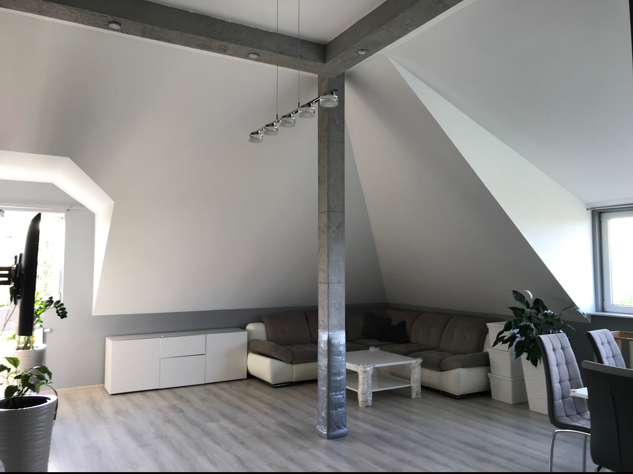 Jasne, przestronne poddasze po remoncie, z szarym narożnikiem, białymi meblami i wykończonymi ścianami, oświetlone nowoczesną lampą wiszącą.