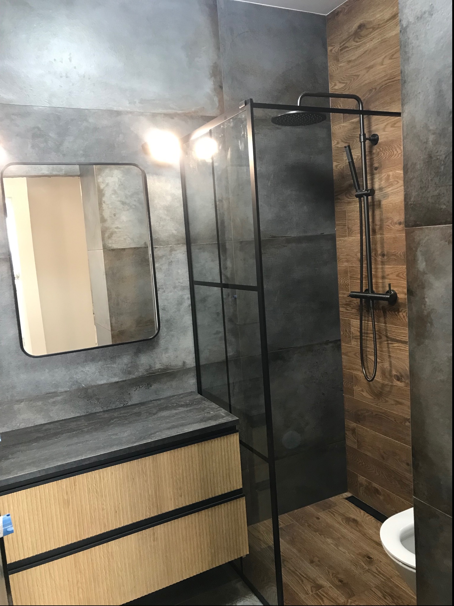 Nowoczesna łazienka z kabiną prysznicową typu walk-in, szarymi płytkami imitującymi beton, drewnopodobnymi płytkami na ścianie, czarną armaturą i szafką pod umywalką z drewnianymi frontami.