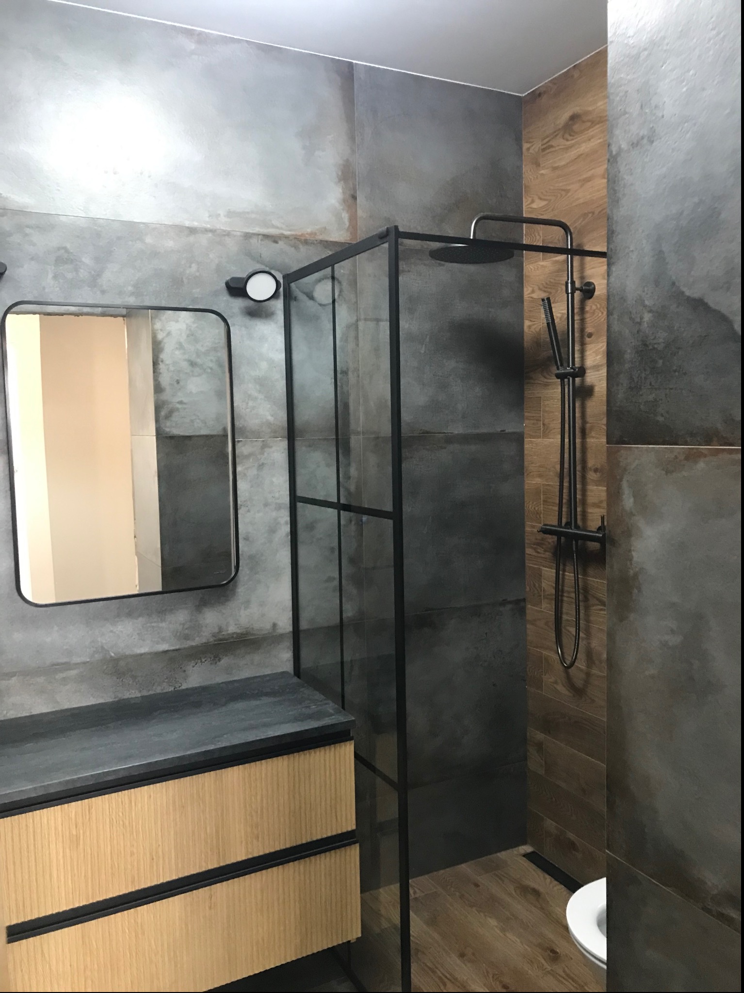 Nowoczesna łazienka z kabiną prysznicową typu walk-in z czarnymi profilami, umywalką z szafką z drewnianymi frontami i lustrem w czarnej ramie na ścianie wykończonej płytkami imitującymi beton.