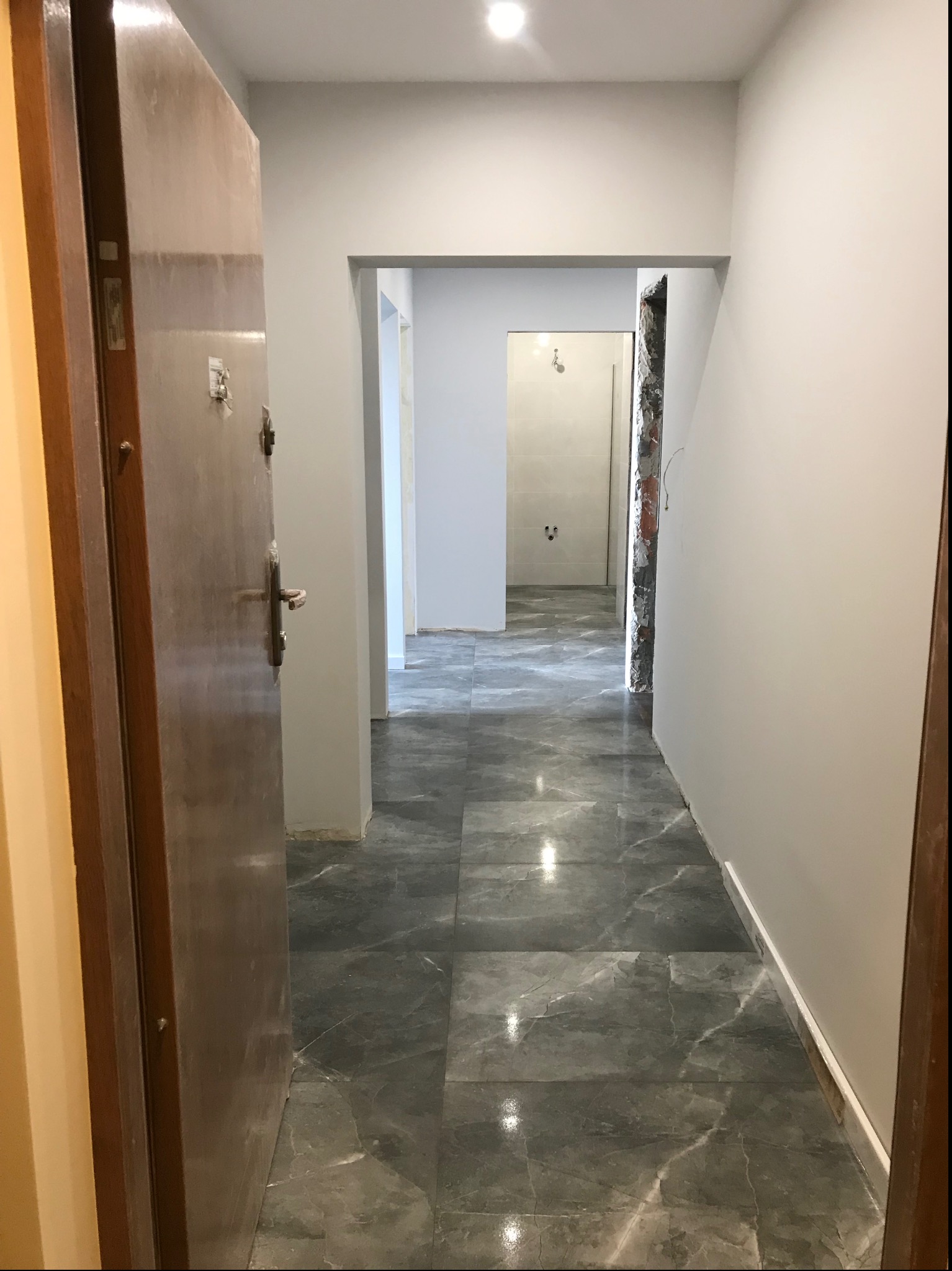 Korytarz po remoncie z szarymi płytkami imitującymi marmur na podłodze, świeżo pomalowanymi ścianami na biało i widocznym wejściem do łazienki w trakcie prac wykończeniowych.